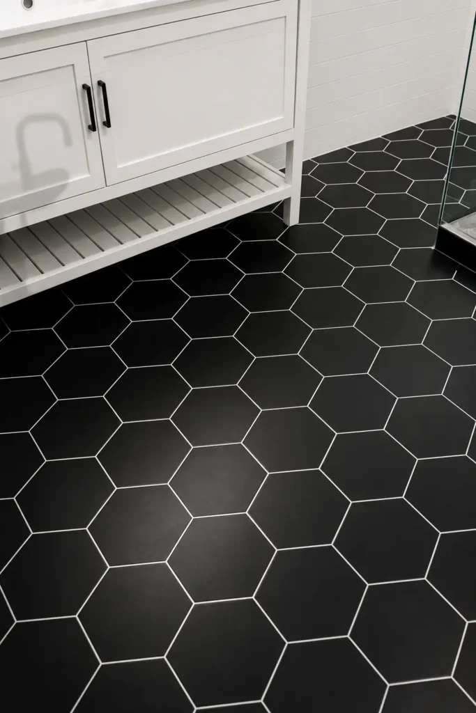 Close-up of a bathroom floor with large black hexagon tiles, crisp white grout lines, a partial white vanity, black cabinet pulls, and a glass shower edge.