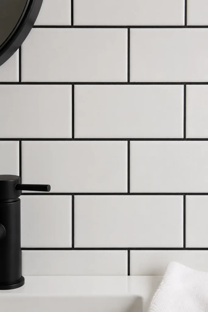 Close-up of white subway bathroom tiles with bold black grout lines, a partial matte black faucet, black mirror edge, and soft white towel detail.